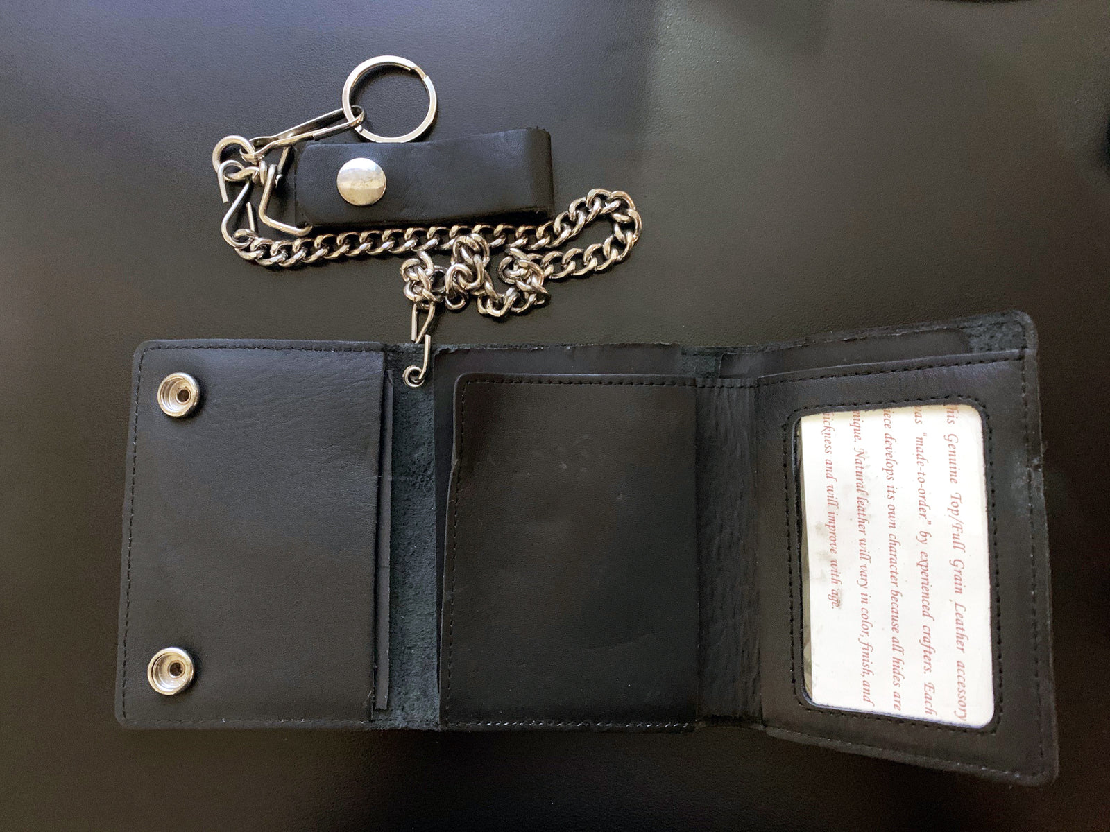 A Cock Sparrer black leather biker wallet with snap buttons is open on a dark surface. Next to it lies a chain with a leather strap and keyring. The wallet features an ID window displaying partially visible text.