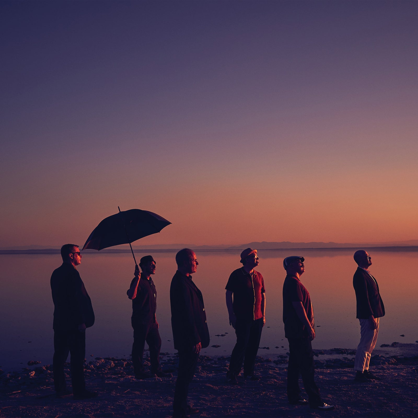 Six people stand in a line on the shore at sunset, one holding an umbrella. Warm purple and orange hues fill the calm, reflective scene—like cover art for The Slackers’ next album.