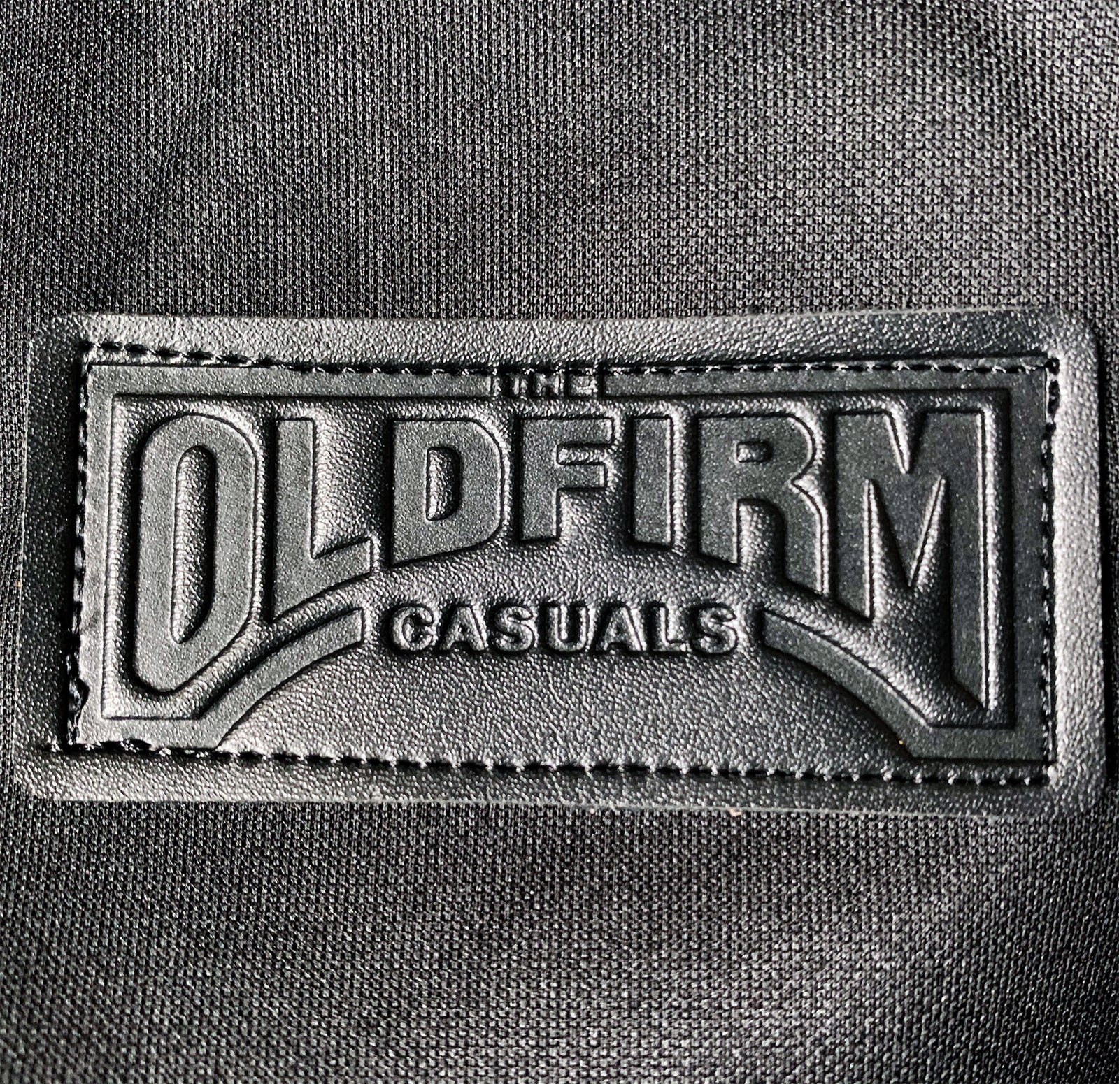 A black leather patch stitched onto fabric, embossed with The Old Firm Casuals in bold capitals—a standout detail on this track jacket.