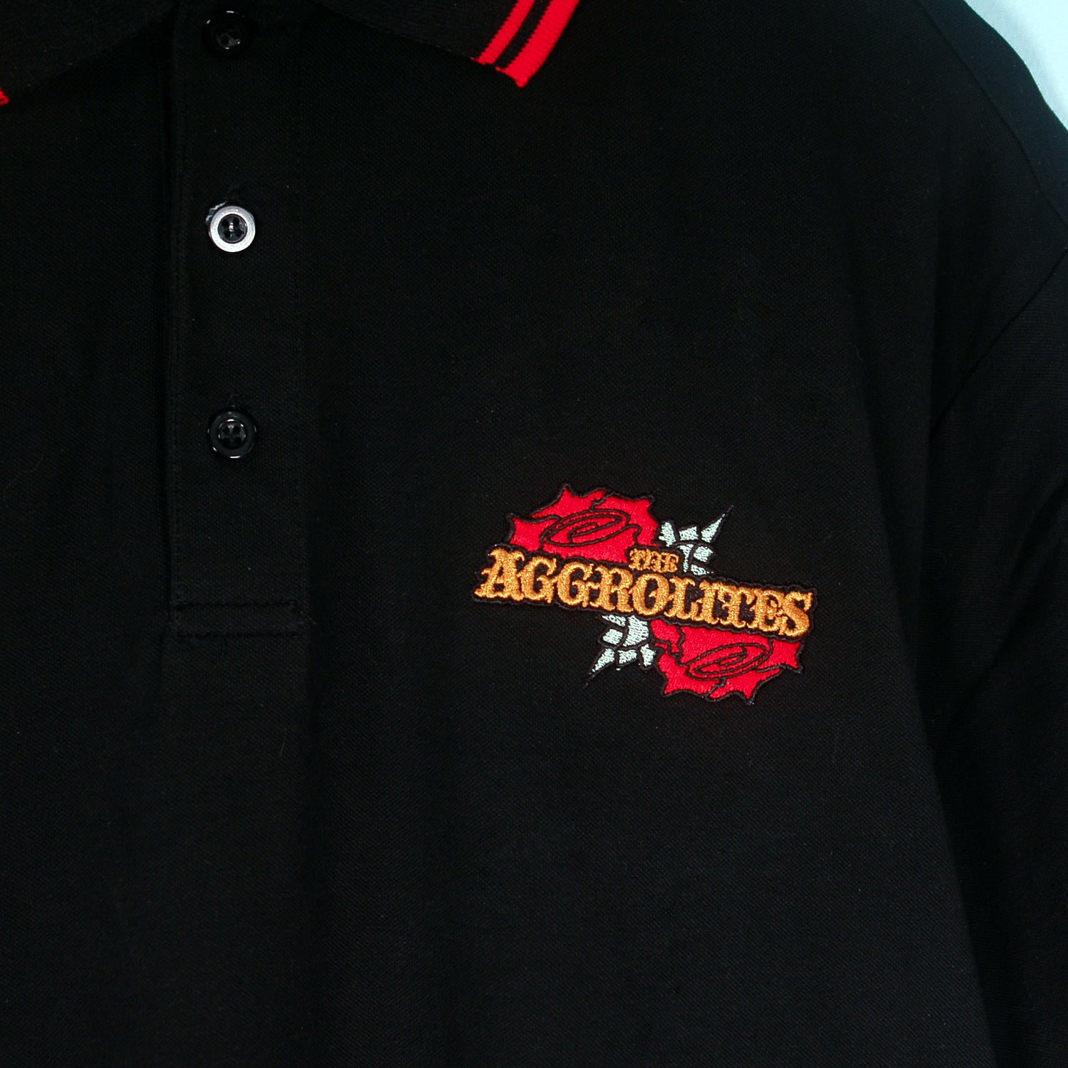 A black The Aggrolites polo shirt featuring red collar trim and an embroidered orange The Aggrolites logo on the chest, surrounded by red roses and white leaves.