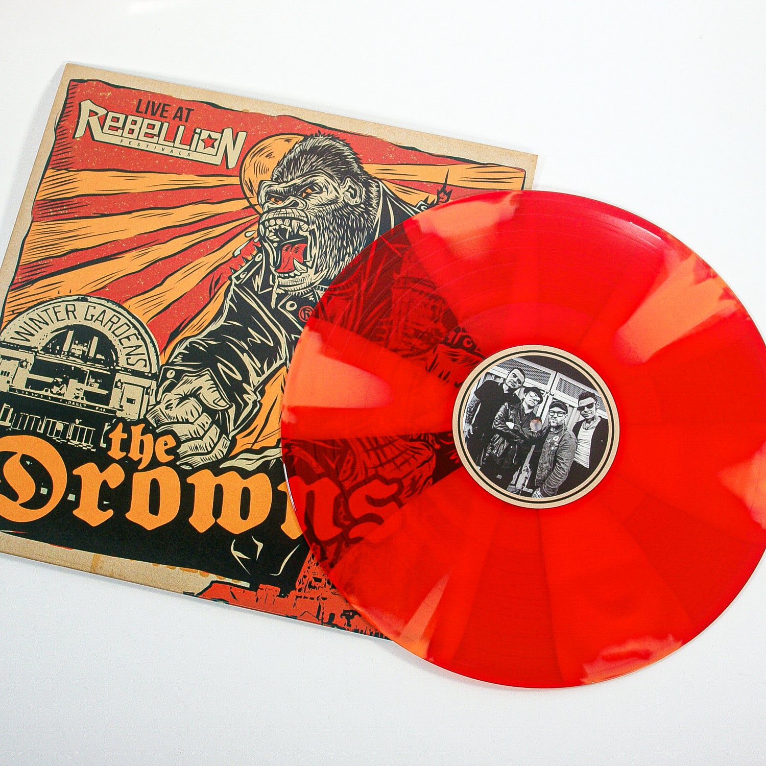 A red and orange marbled vinyl from The Drowns sits beside its cover, which shows a roaring gorilla in a suit destroying buildings, with The Drowns and Live at Rebellion prominently displayed.