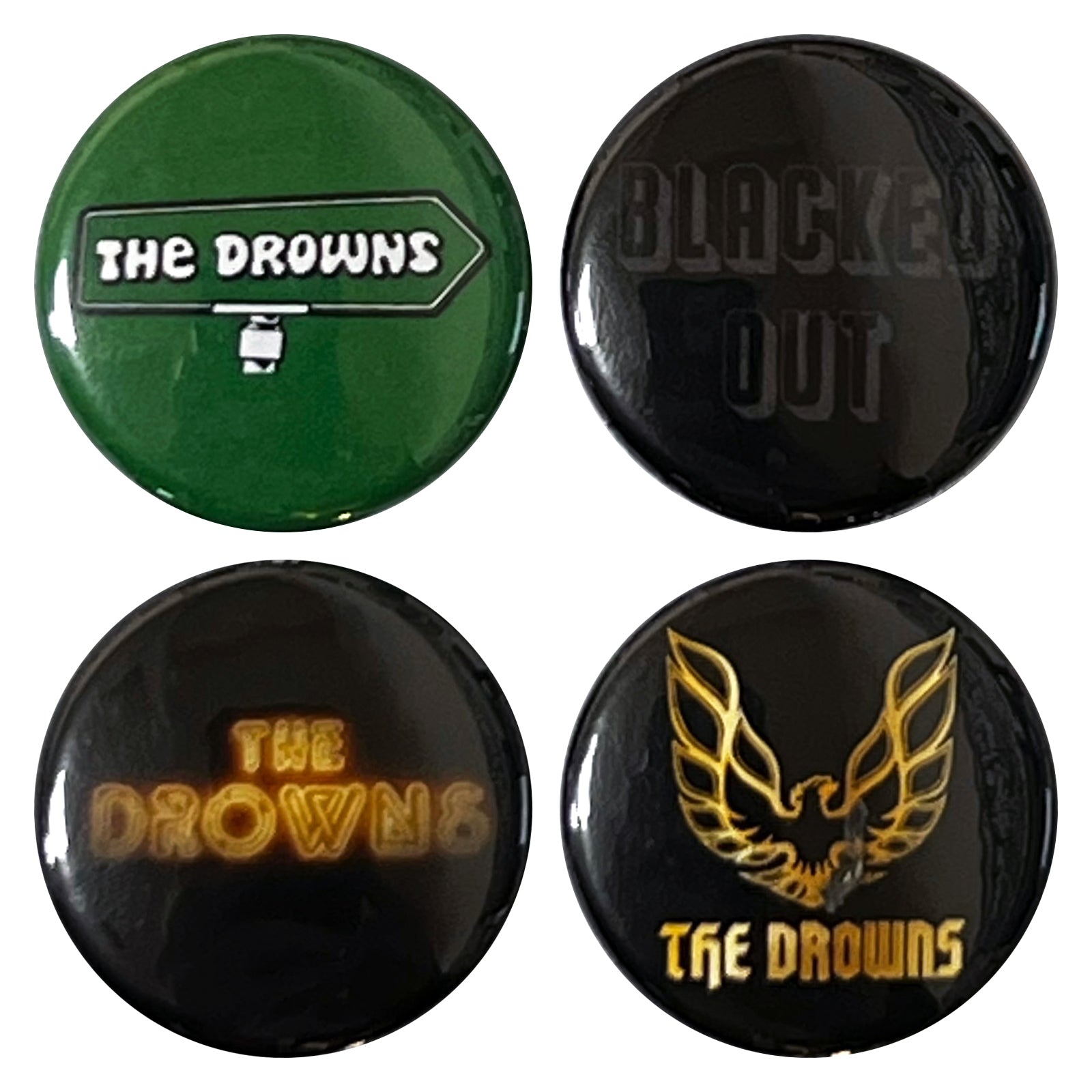 This The Drowns button pack includes four round pins on a hang card: green with THE DROWNS and sign, blacked out with BLACKED OUT in dark letters, black with THE DROWNS in yellow, and one gold bird design.