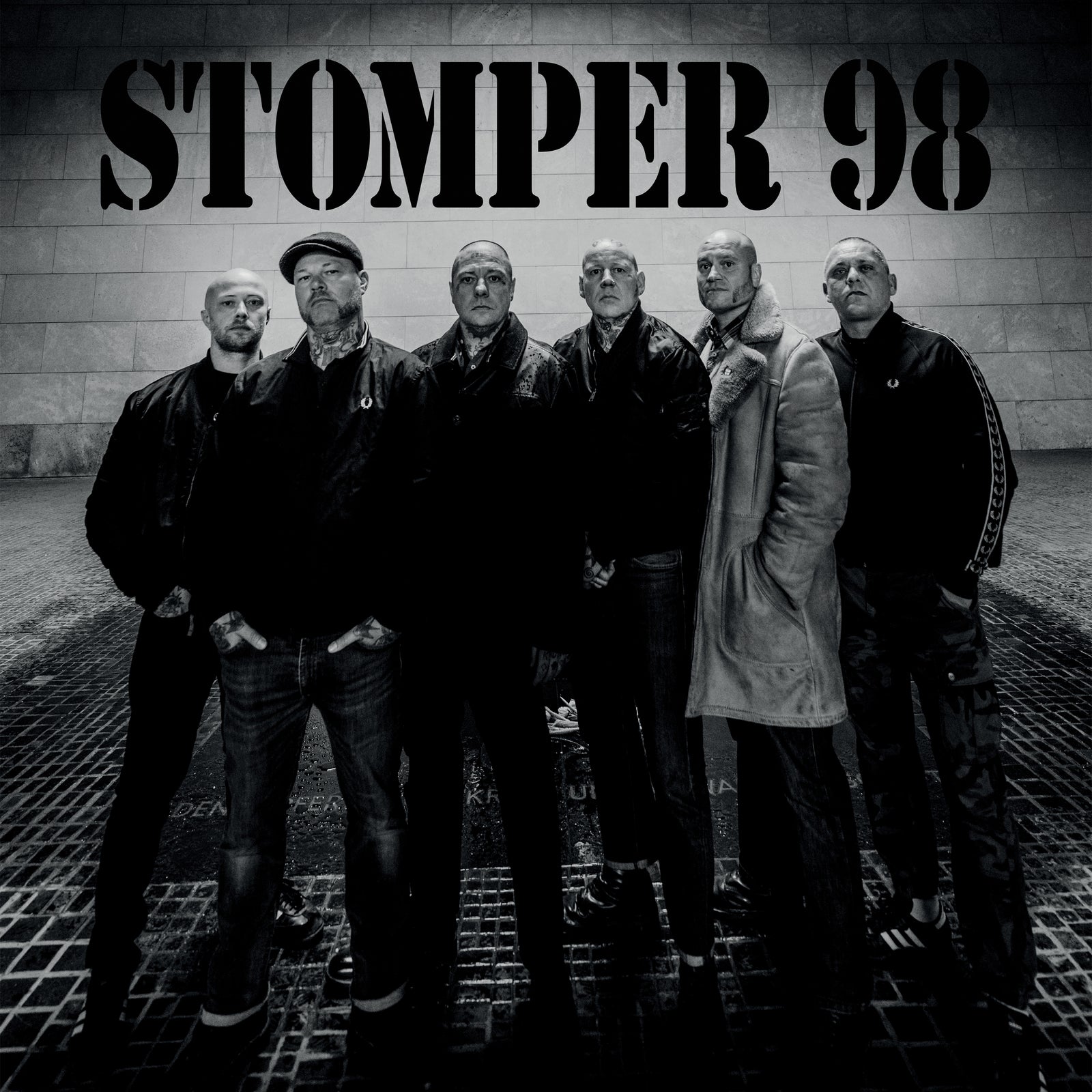 Six men with serious expressions stand in a row under bold Stomper 98 text. This black-and-white image captures the German Oi! band in jackets and coats against a stone wall, showcasing Stomper 98s working-class punk rock spirit.