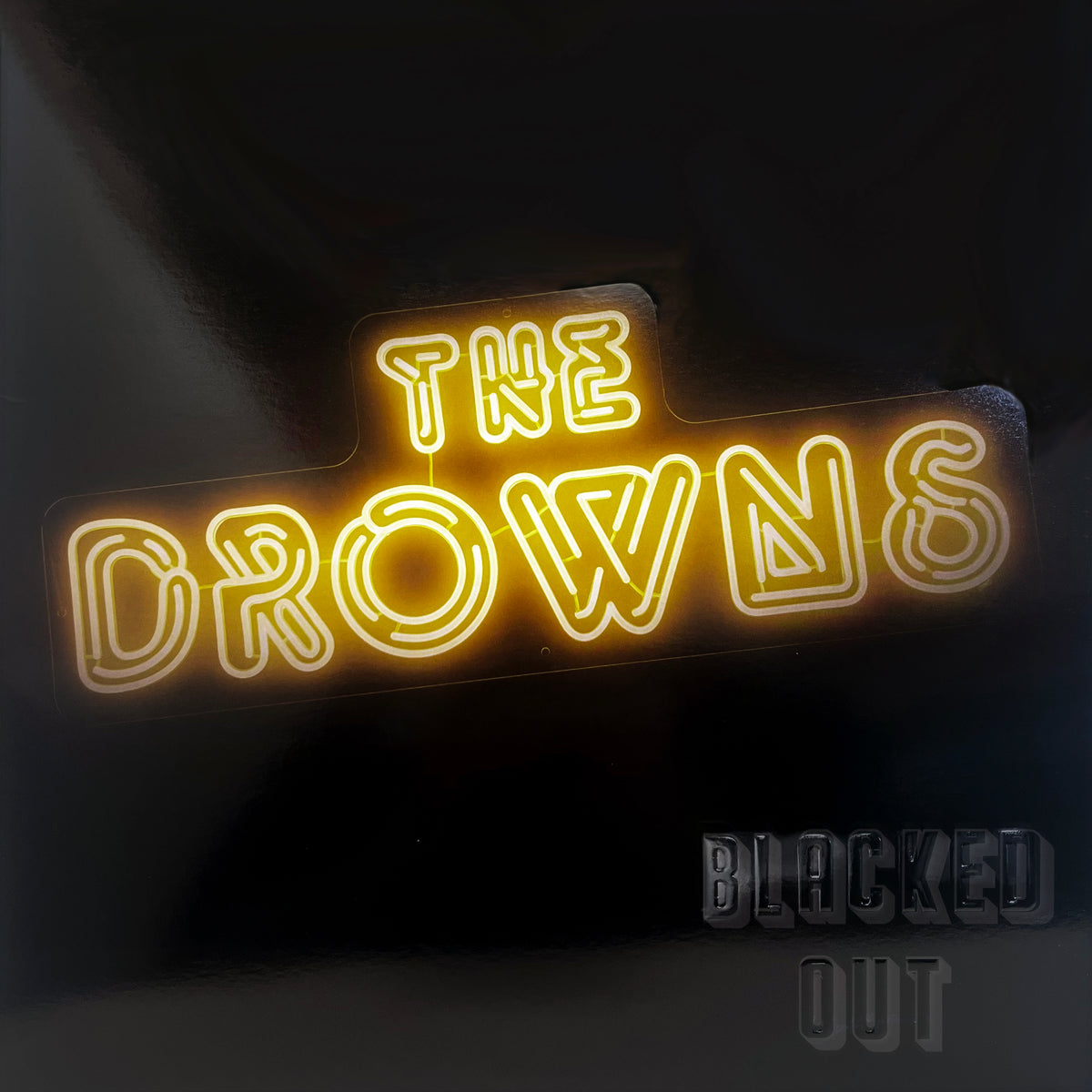 A glowing yellow neon sign displays The Drowns (Brand Name) on a black background, radiating punk rock energy. Blacked Out is written in a faded, dark font in the lower right corner.