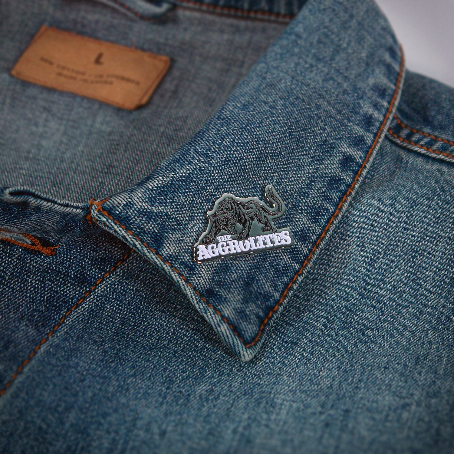 A denim jacket collar displays The Aggrolites enamel pin—a black lion with "THE AGGROLITES" in white—plus a visible brown label inside.