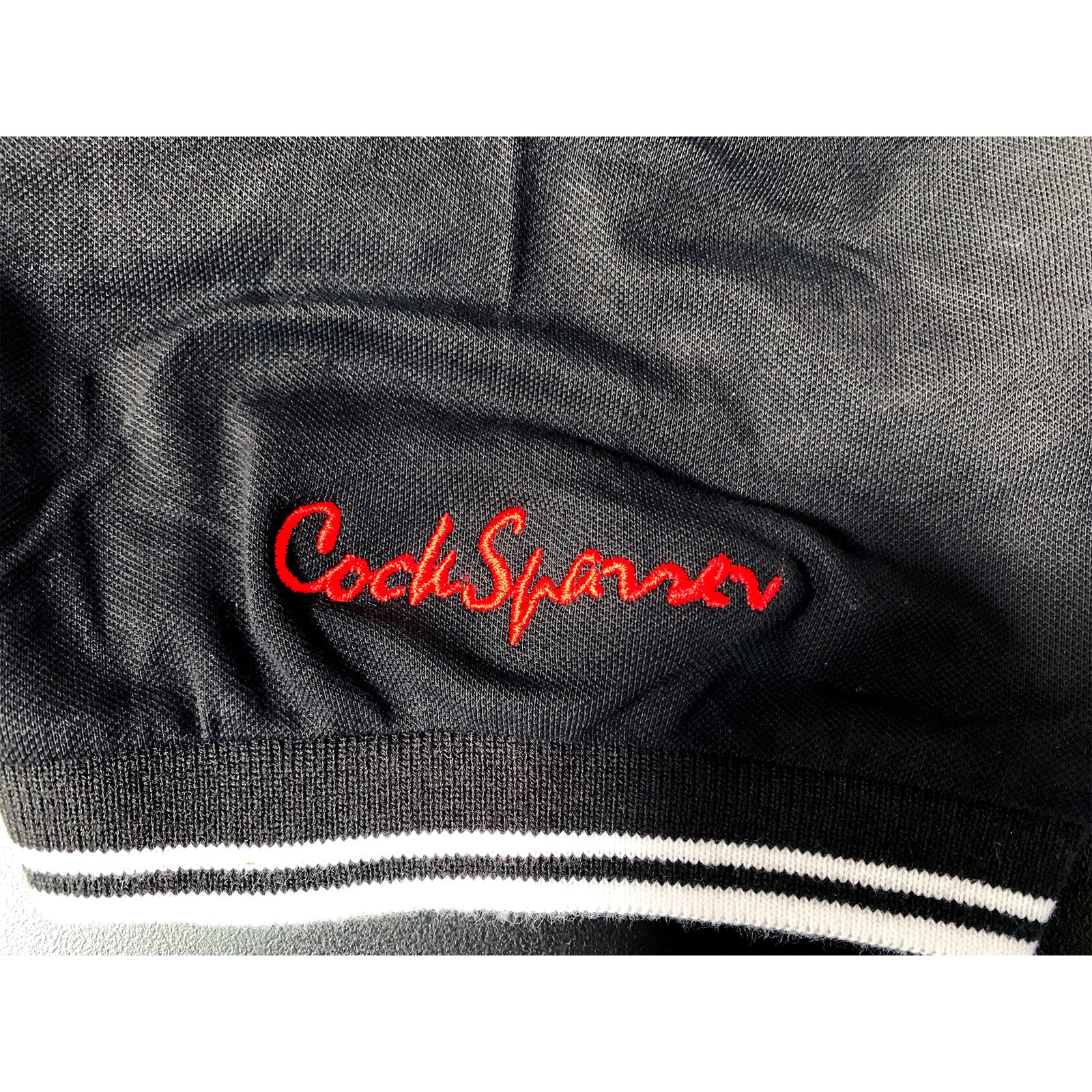Black fabric with Cock Sparrer’s name embroidered in bold red cursive, finished with a black and white striped elastic trim—classic polo-inspired style that pays tribute to the iconic band.