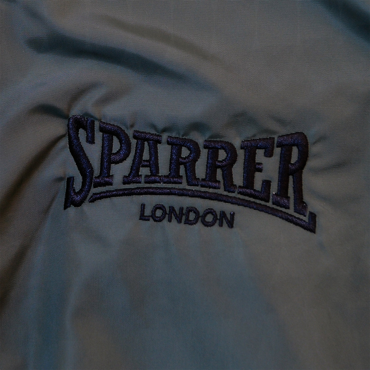 Navy blue embroidery on fabric spells SPARRER LONDON in bold, uppercase letters, matching the Cock Sparrer logo featured on their bomber jacket. The stitching contrasts with the dark, smooth material. Brand: Cock Sparrer.