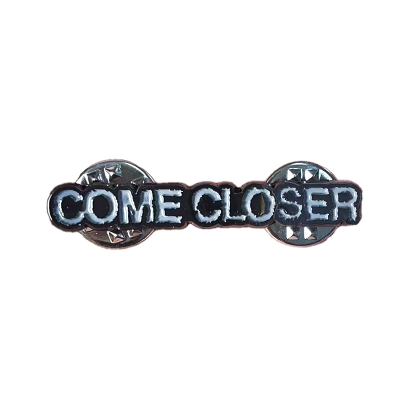 The Come Closer pin is a 1.5-inch black enamel accessory with COME CLOSER in white capitals, silver foil details, and two silver butterfly clutch backings.