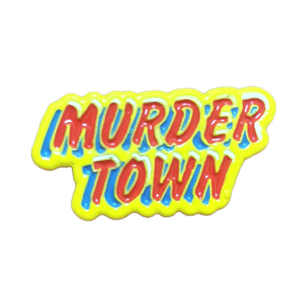 The Grade 2 brand’s striking 1-inch enamel pin displays &quot;MURDER TOWN&quot; in bold red letters outlined in blue and yellow, set on a vivid yellow background.