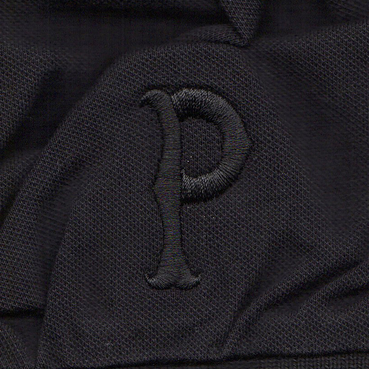 A black polo with a textured finish features a raised, embroidered letter P in black thread—ideal for fans of Pirates Press Records.