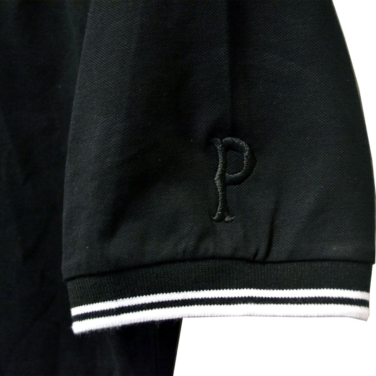 A close-up of a black Pirates Press Records polo featuring white and black striped cuffs and an embroidered letter P on the sleeve, inspired by their iconic One Family One Flag design.