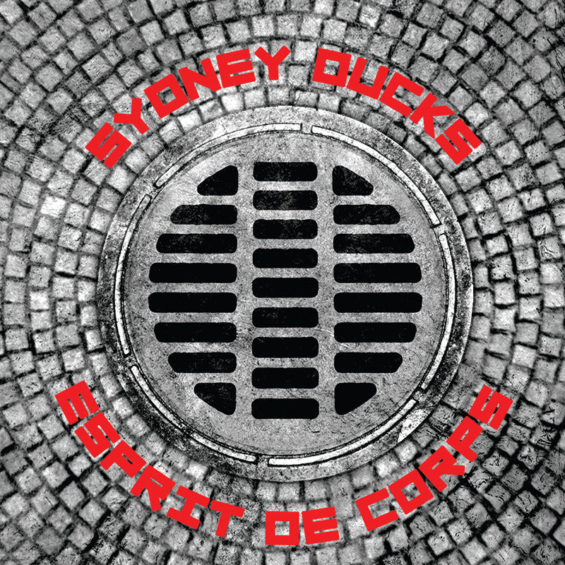 A circular metal grate on cobblestone, with &#39;Sydney Ducks&#39; above and &#39;Esprit de Corps&#39; below in bold red letters, captures the gritty energy of San Francisco&#39;s punk-inspired brand, Sydney Ducks.