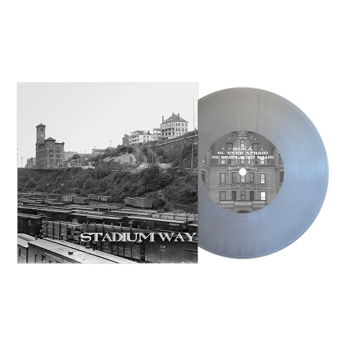 A translucent gray 7″ vinyl record sits next to an album cover featuring a black-and-white photo of historic buildings, railroad tracks, and boxcars, with the text STADIUM WAY across the lower part of the cover—an evocative release from this acoustic project.