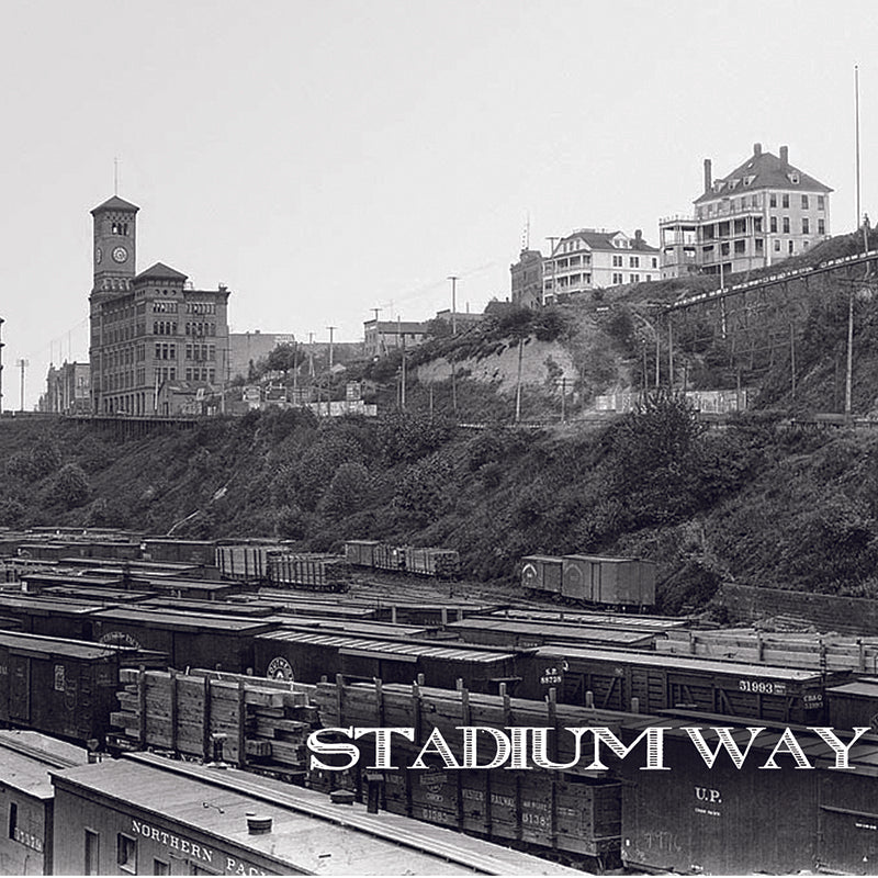 Black and white photo of train cars on tracks below a hill, historic buildings and a clock tower above. Large Stadium Way text at the bottom—ideal for an acoustic project or 7 vinyl cover. Brand: Stadium Way.