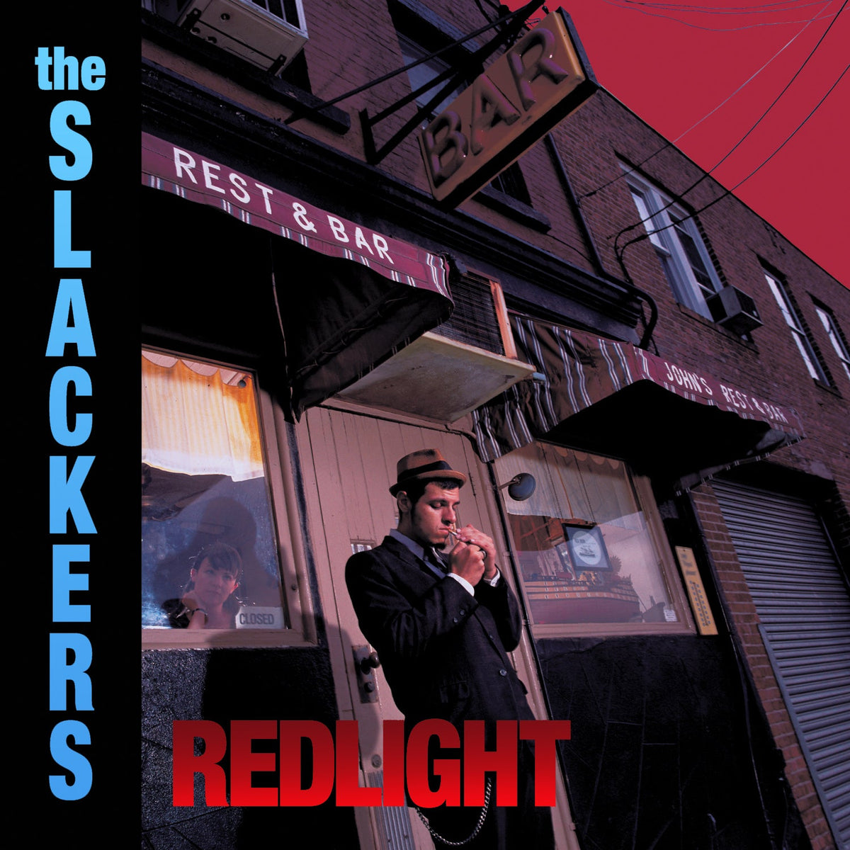 A man in a suit and hat lights a cigarette outside a bar with striped awnings at dusk. The Slackers album title appears on the left, while “REDLIGHT” in red at the bottom refers to the sought-after Redlight vinyl by The Slackers.