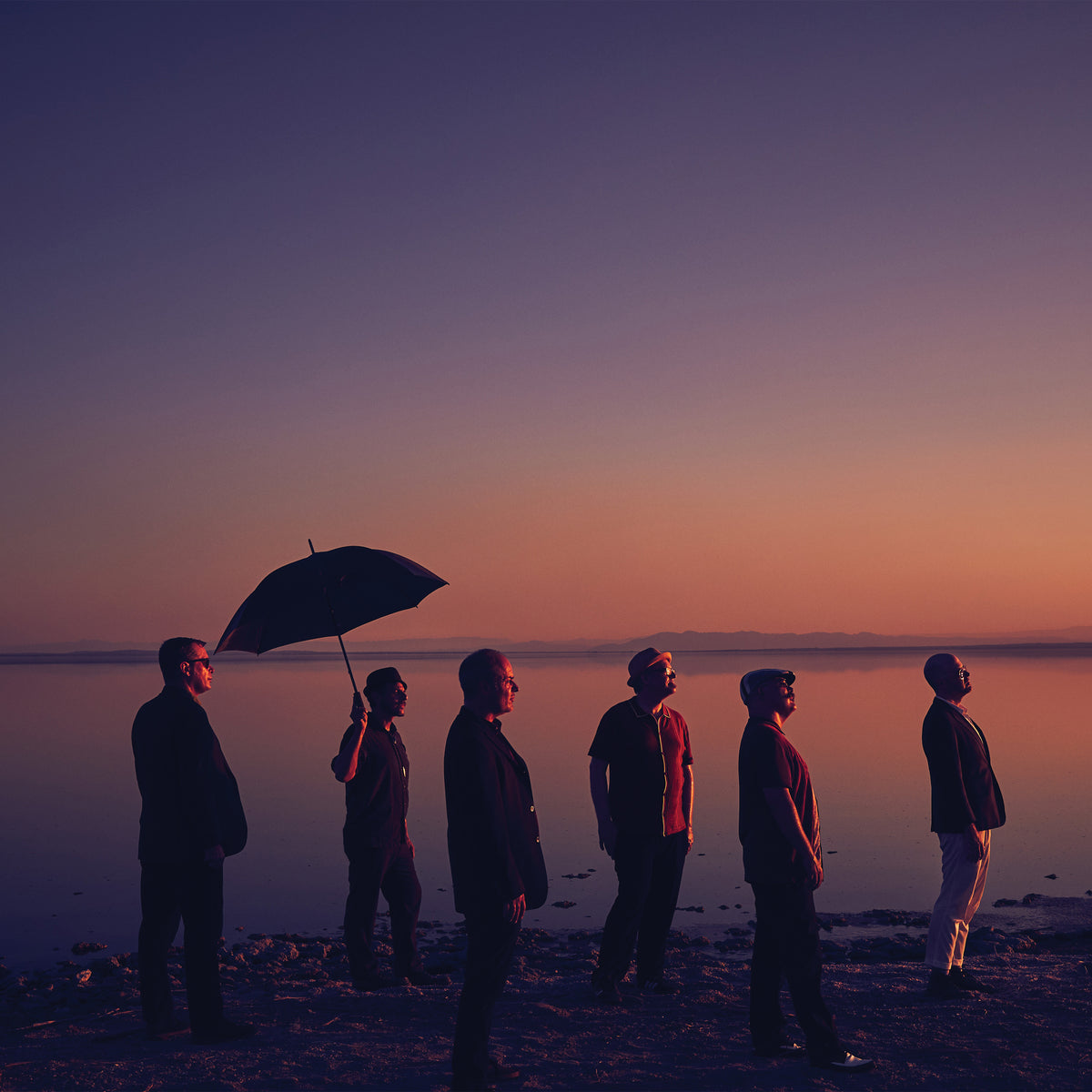 Six people stand by calm water at sunset, one with an umbrella. Their silhouettes and long shadows evoke the serene, contemplative mood of The Slackers new album from ska band The Slackers.