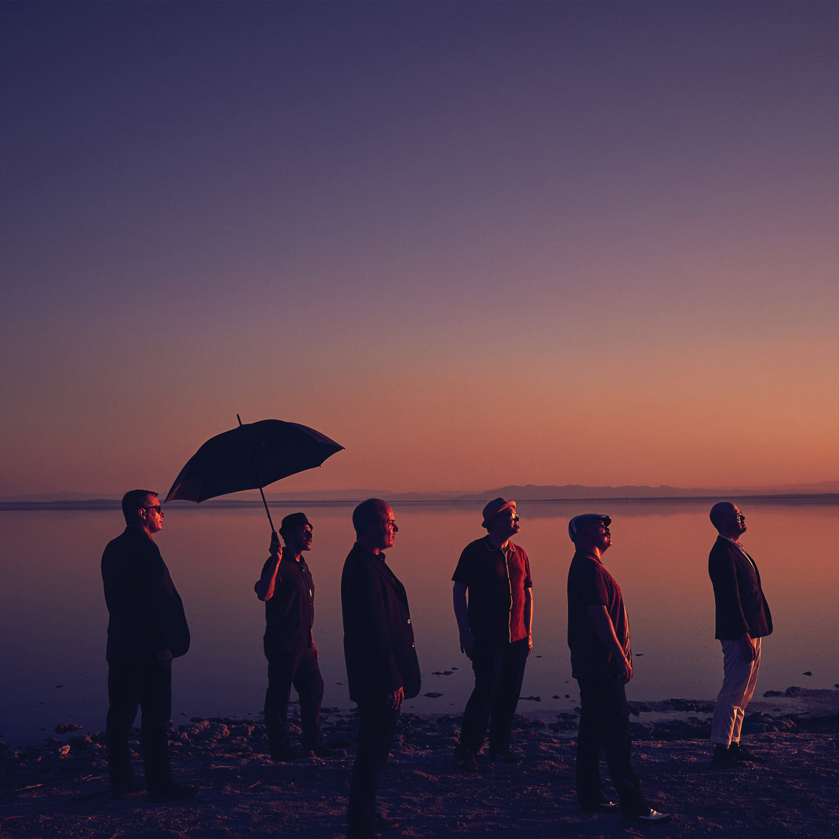 Six people stand in a line on the shore at sunset, one holding an umbrella. Warm purple and orange hues fill the calm, reflective scene—like cover art for The Slackers’ next album.