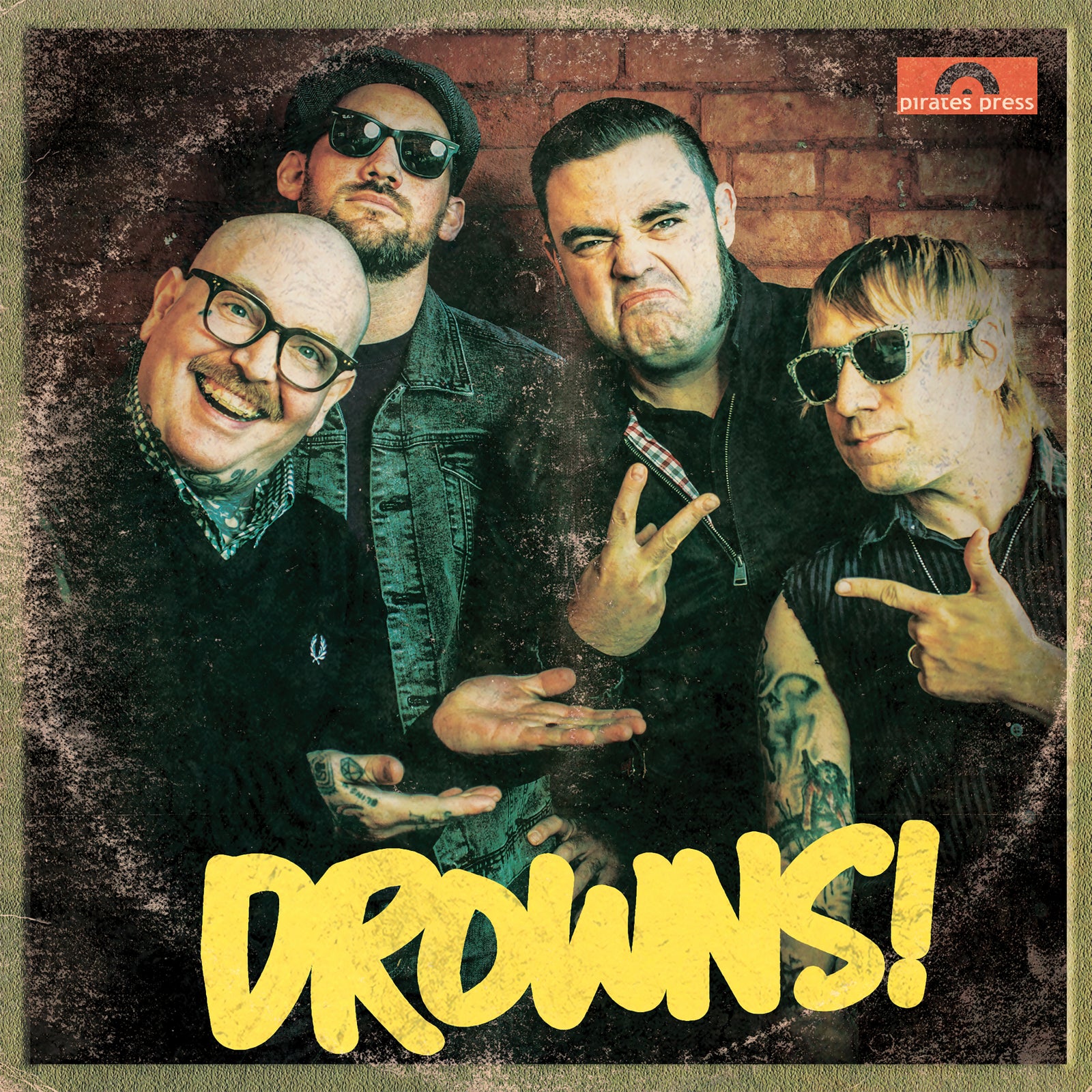 Four men pose before a brick wall, striking playful or tough looks; three wear sunglasses, one wears glasses. DROWNS! is boldly displayed in yellow below, capturing The Drowns’ classic rock and roll energy. Brand: The Drowns.