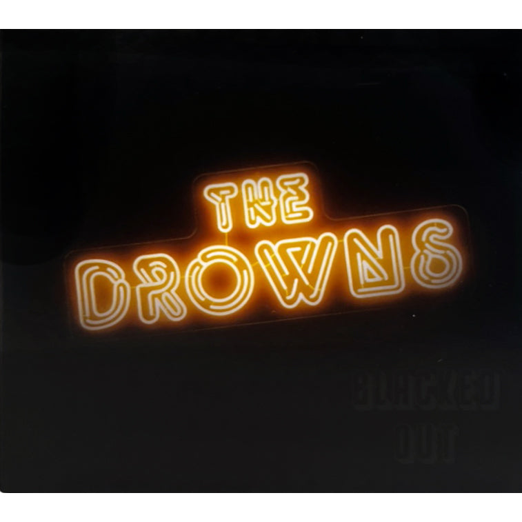 A glowing neon sign reads The Drowns in orange-yellow letters on a black background, capturing their blue-collar punk rock energy. Blacked Out is faintly visible in the bottom right corner.