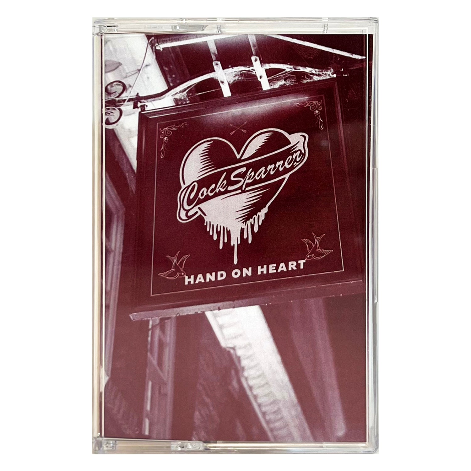 A sepia-toned photo shows a sign with a heart and the words Cock Sparrer and Hand On Heart, embodying punk rock spirit as it hangs above a street, with part of a building and ceiling in the background. Brand: Cock Sparrer.