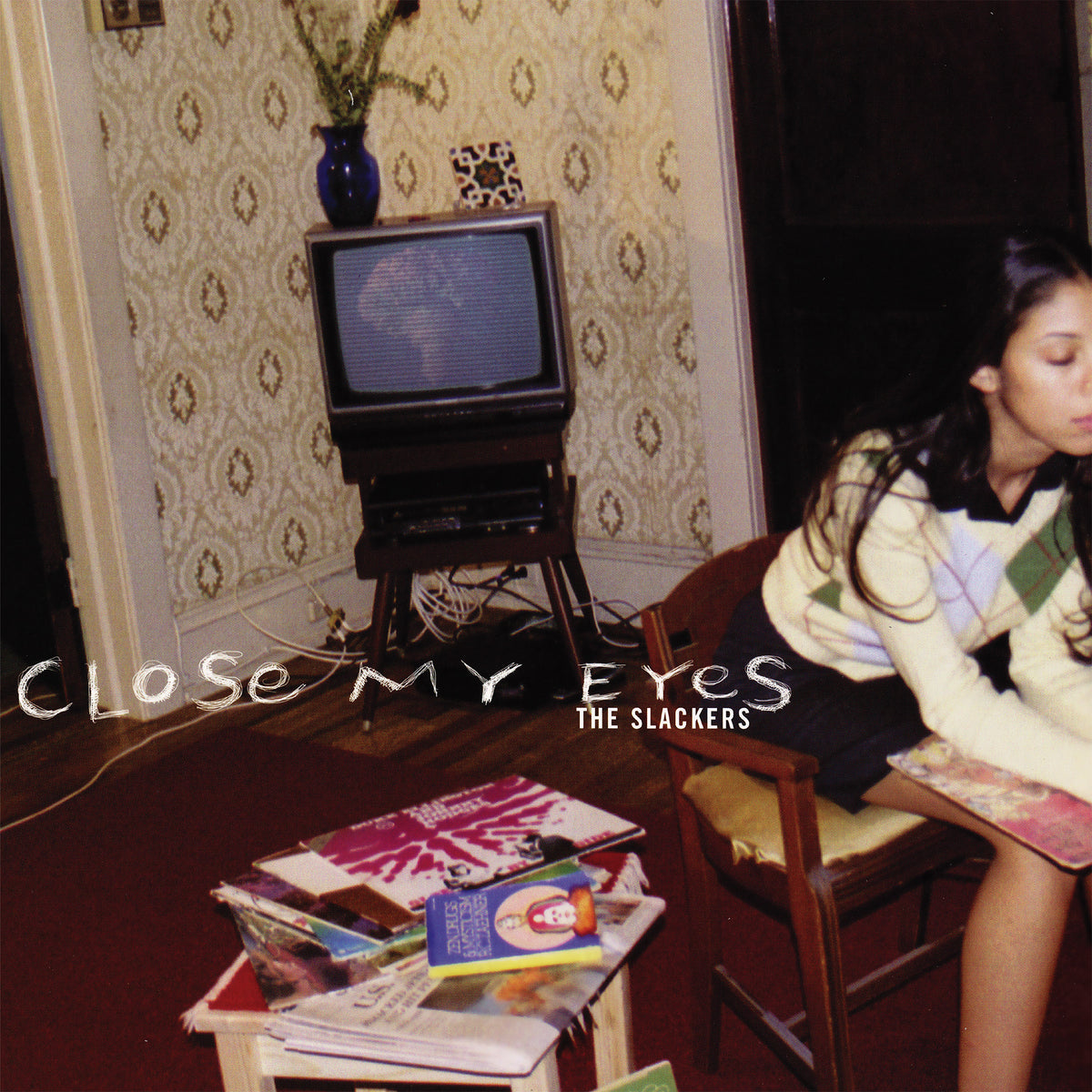 A young woman sits in a retro living room with books and magazines, an old TV, and patterned wallpaper, capturing NYC ska-reggae vibes. Text reads: Close My Eyes by The Slackers.