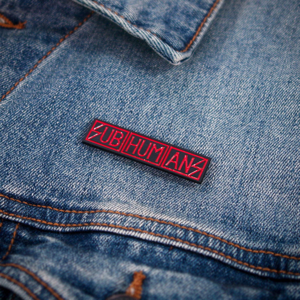 A Subhumans rectangular pin with the band’s name in bold red letters on a black background is attached to the pocket of a blue denim jacket.