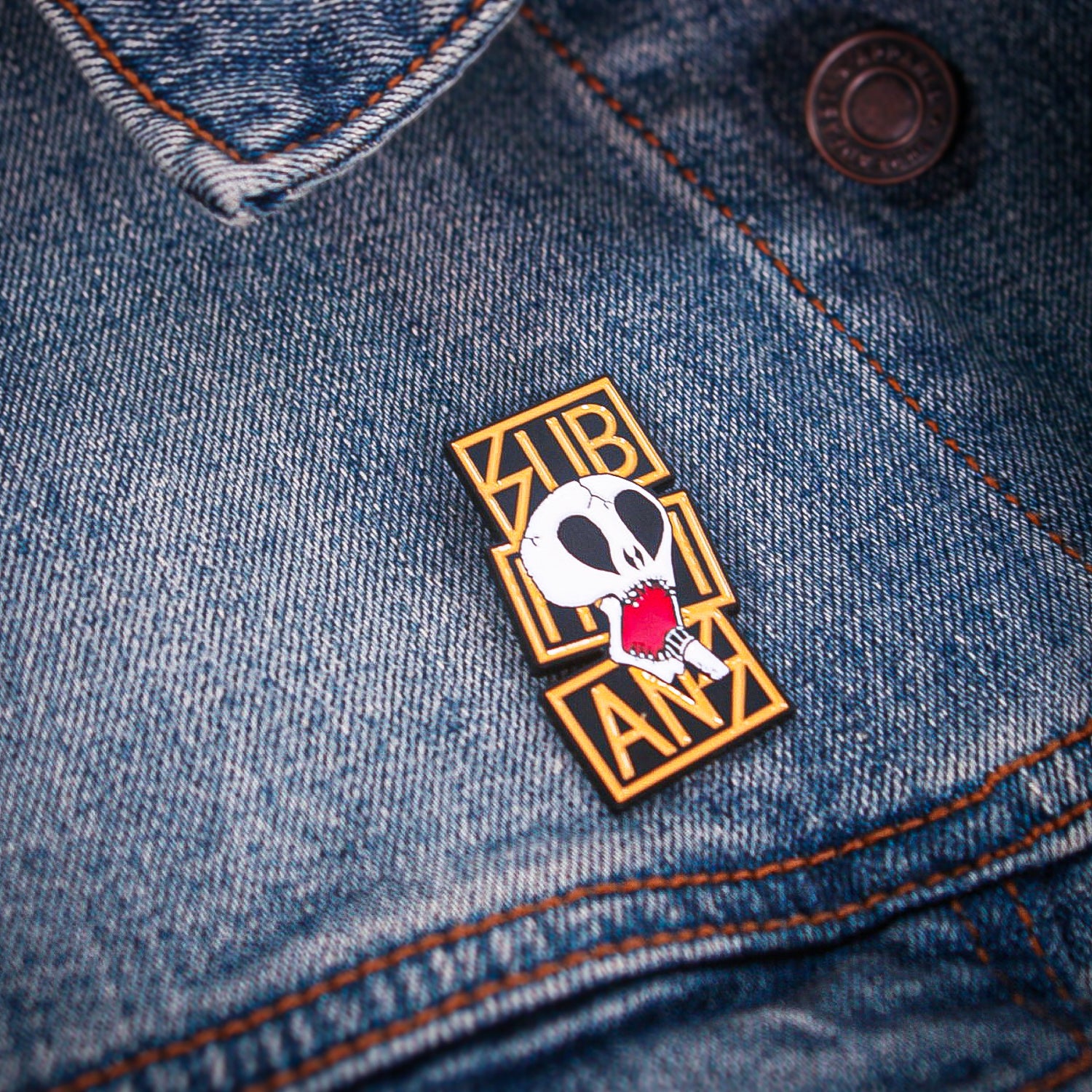 A gold and black enamel pin by Subhumans features a stylized skull with open mouth and the word SUBLIMINAL in bold stacked letters, shown on blue denim near a jacket button.