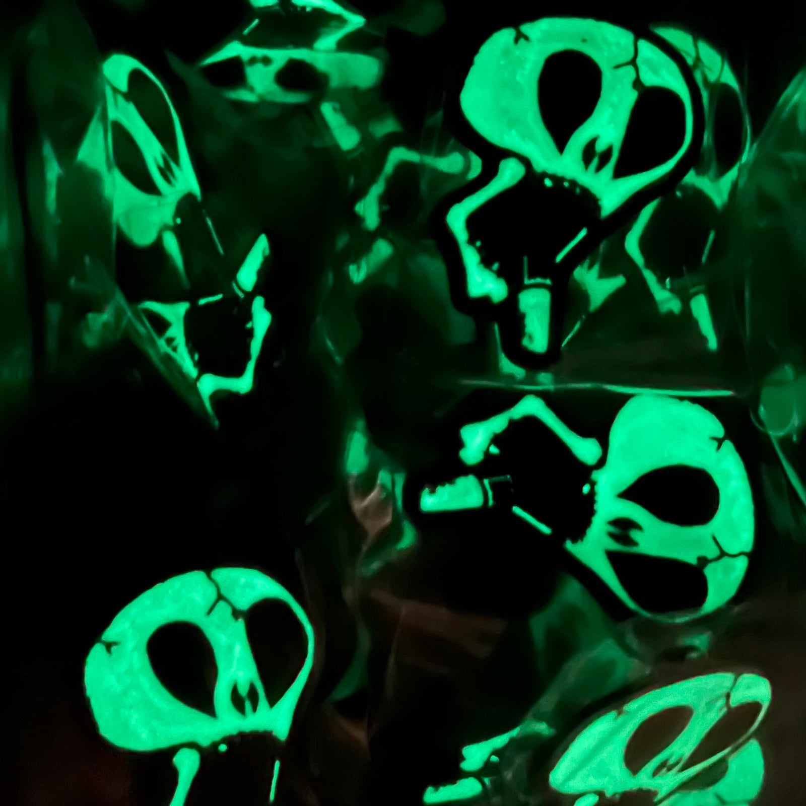 Subhumans glowing green skull-shaped enamel pins with large black eye sockets are scattered on a dark background, creating an eerie, otherworldly glow-in-the-dark effect.