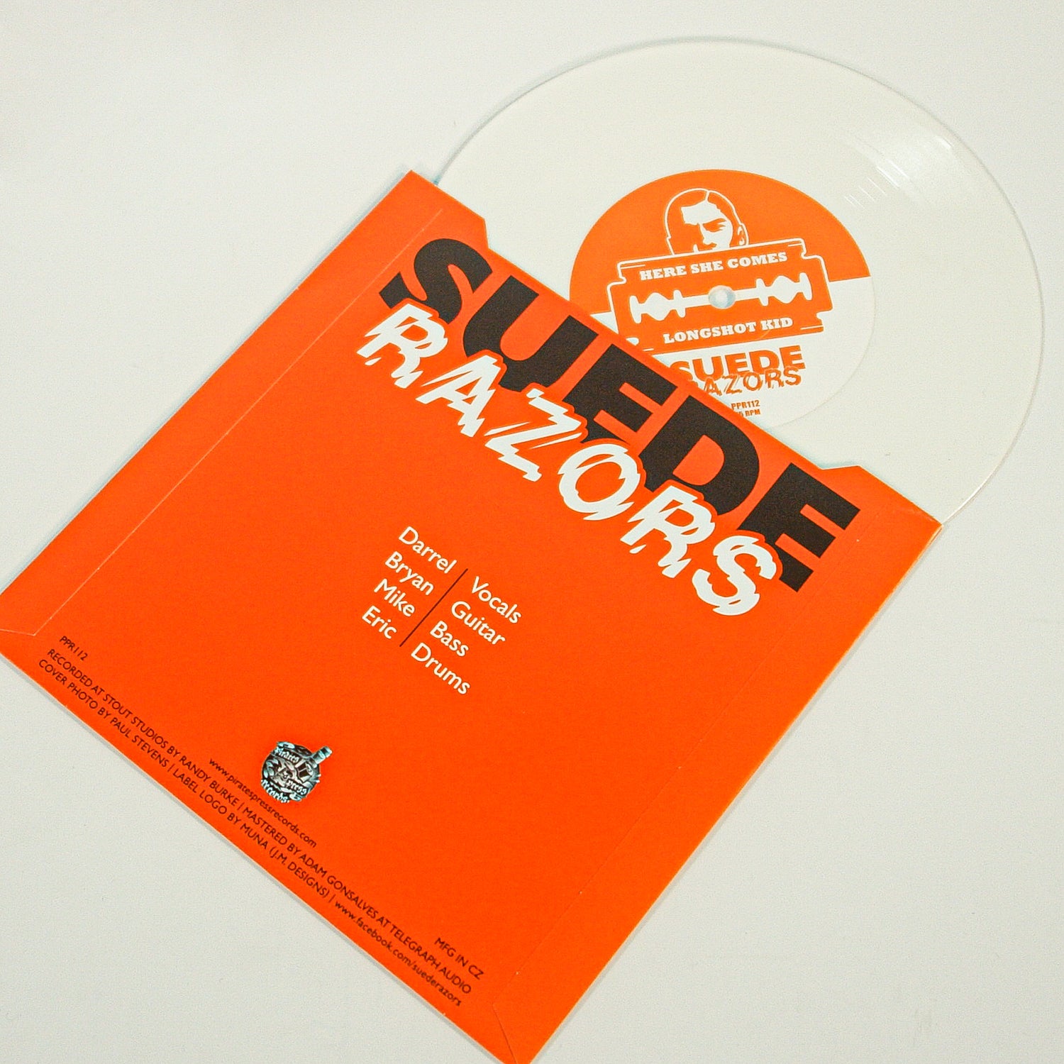 A clear vinyl record partly out of an orange Suede Razors sleeve lists band members and roles. The label features Here She Comes by Suede Razors in retro orange and white rock 'n' roll text.