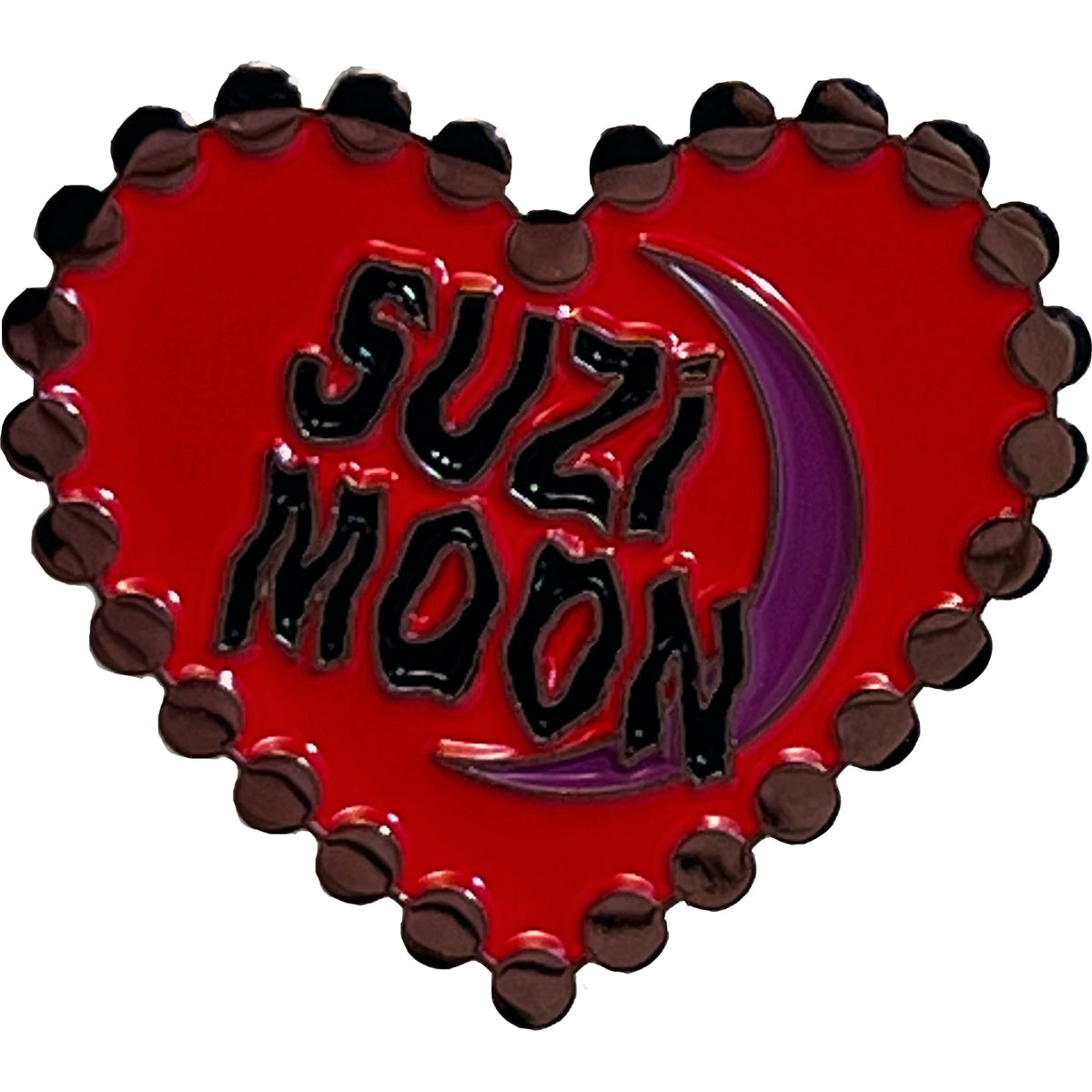 A red, heart-shaped Suzi Moon enamel pin with a black scalloped border showcases bold black &quot;SUZI MOON&quot; text and a purple crescent moon on the right.