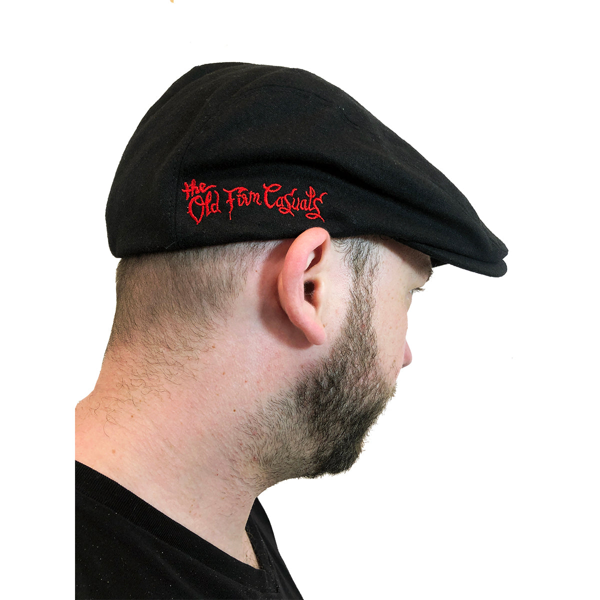 A bearded man wears a black scally cap from The Old Firm Casuals featuring red embroidered text on the side, set against a white background.