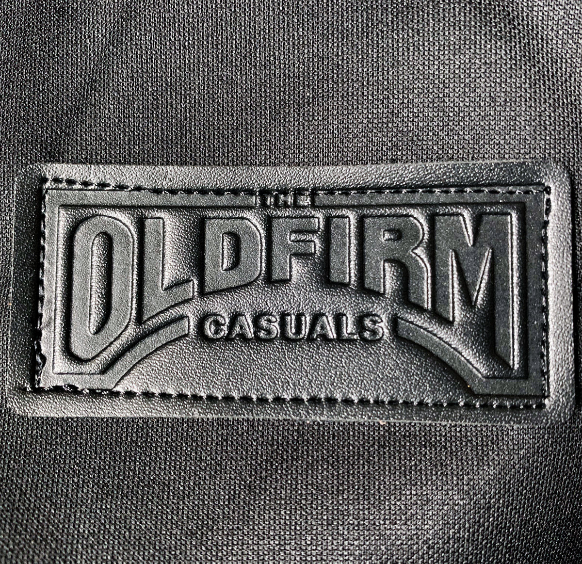 A black leather patch stitched onto fabric, embossed with The Old Firm Casuals in bold capitals—a standout detail on this track jacket.