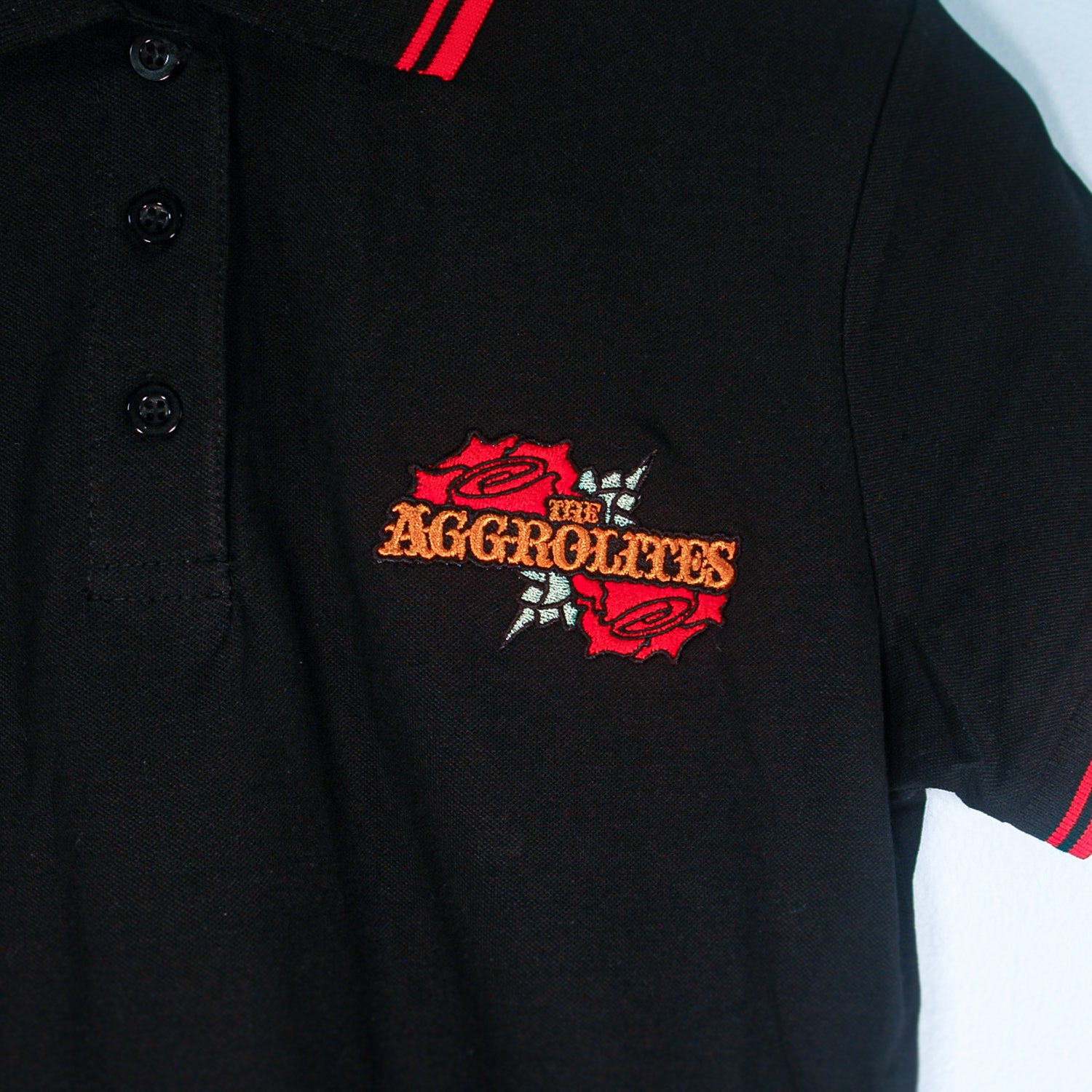 A black The Aggrolites polo shirt with red trim on the collar and sleeves, featuring an embroidered logo of two red roses around orange and yellow The Aggrolites text.