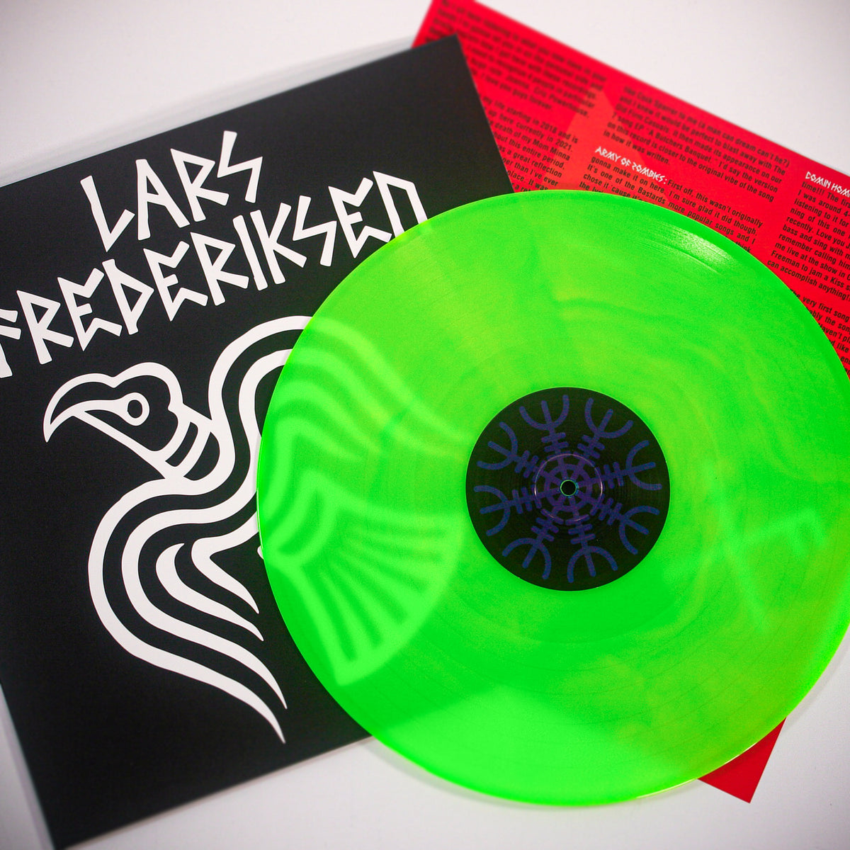 A Neon Green Vinyl record partly covers an album sleeve with bold white Lars Frederiksen text and a bird graphic. Behind them, a red lyrics sheet hints at tracks from the album To Victory.