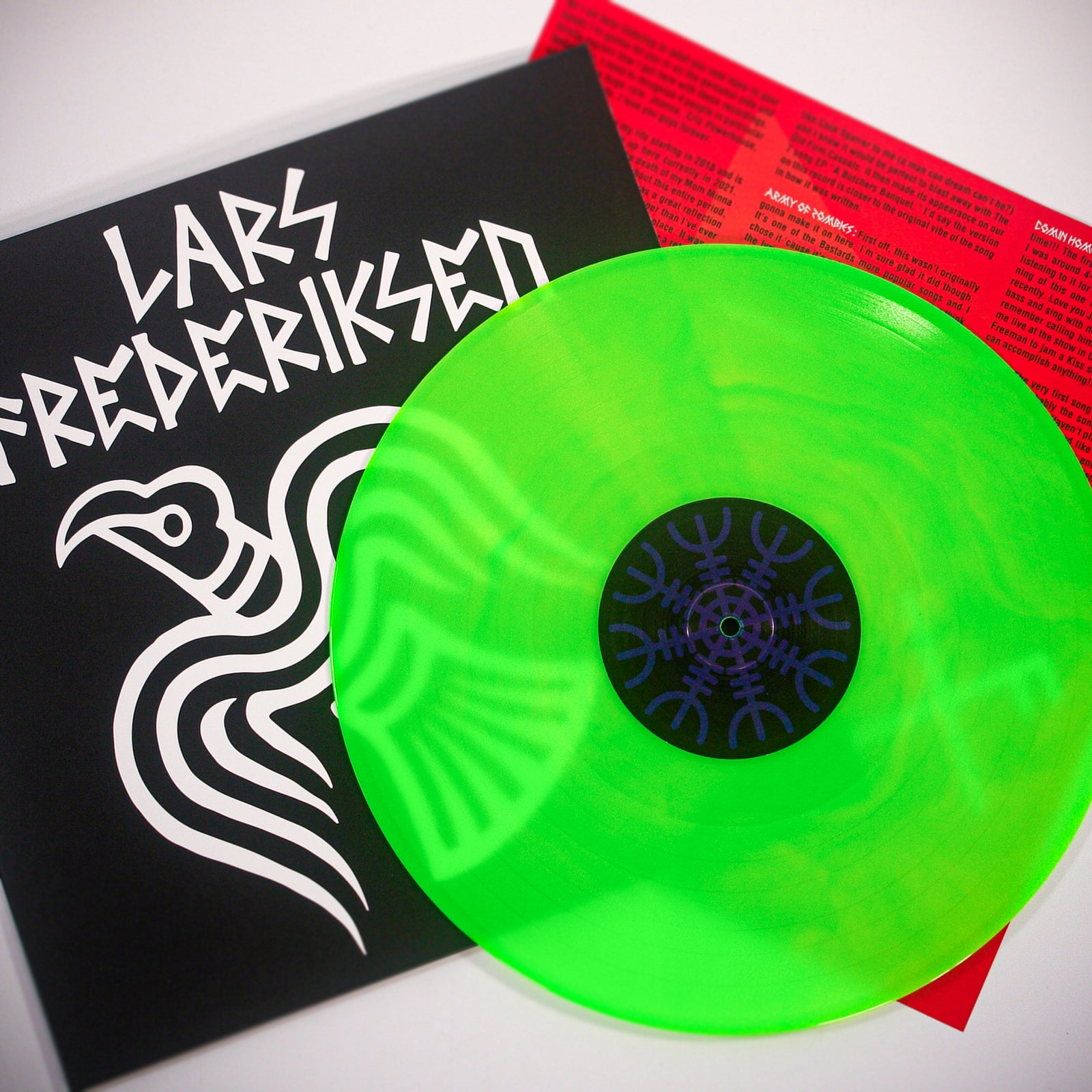 A Neon Green Vinyl record partly covers an album sleeve with bold white Lars Frederiksen text and a bird graphic. Behind them, a red lyrics sheet hints at tracks from the album To Victory.
