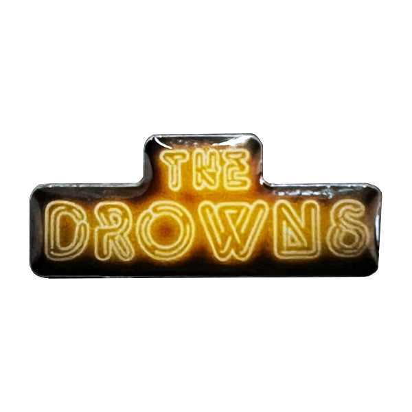 A 1-inch enamel pin by The Drowns, featuring bold, yellow neon-style lettering on a dark background.