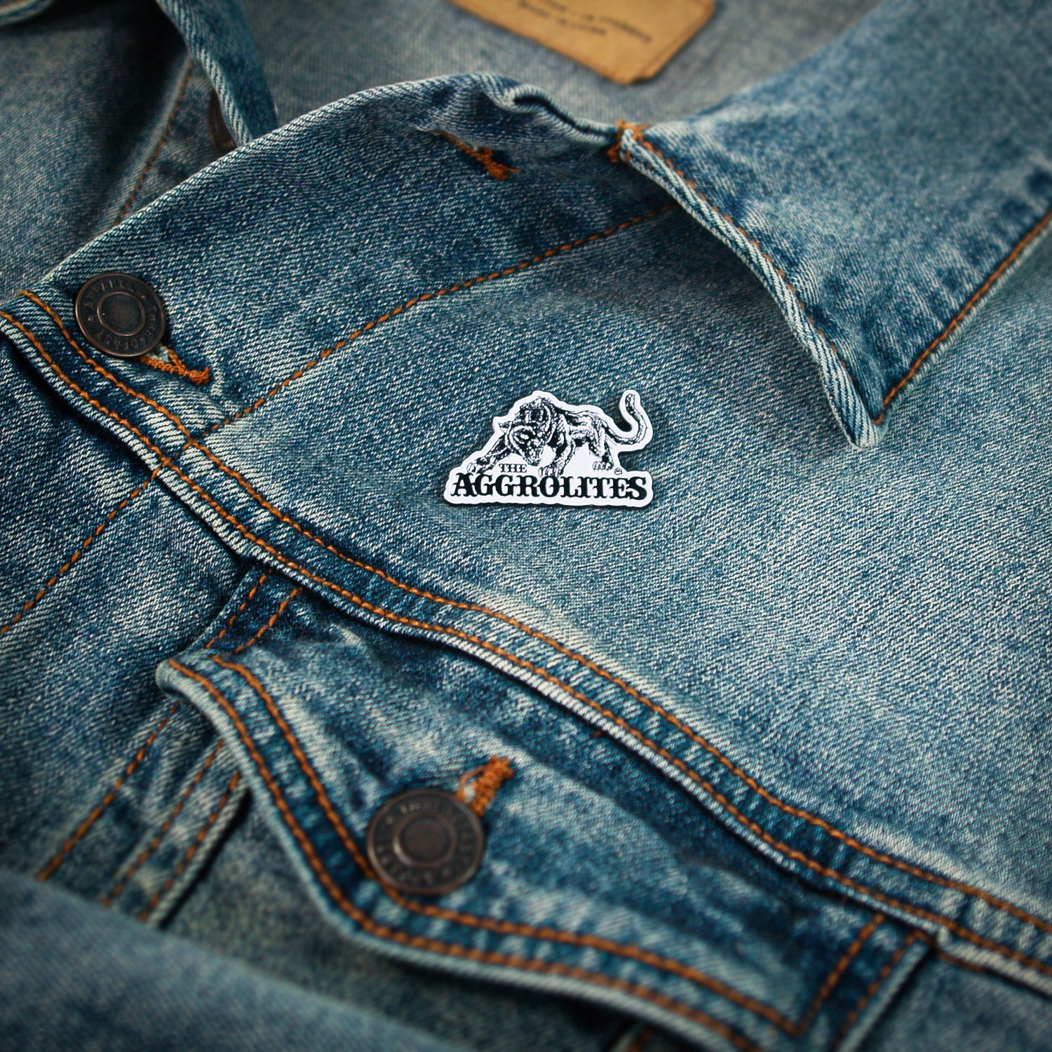 A close-up of a blue denim jacket shows a black and white enamel pin on the pocket flap. The pin features The Aggrolites’ Aggropanther design and the band's name.