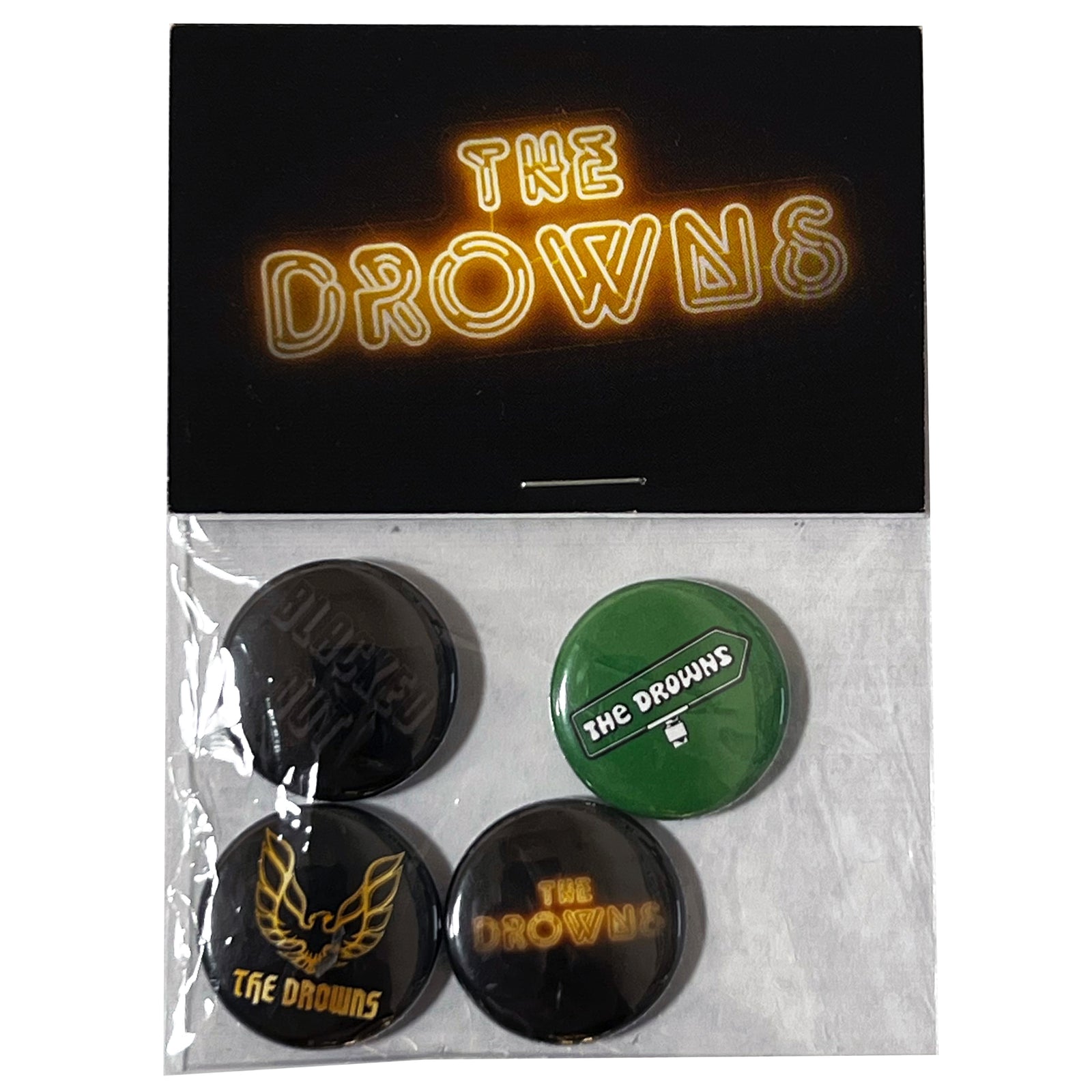 A set of four The Drowns band buttons in a plastic bag, displayed on a hang card with The Drowns in yellow neon-style text. Includes assorted designs such as blacked-out buttons and classic band logos. Brand: The Drowns.