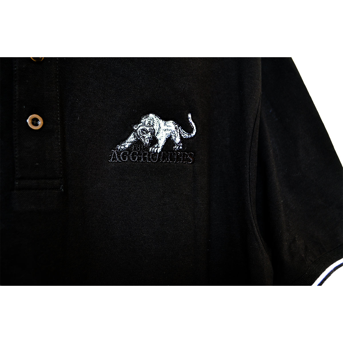 Close-up of a black The Aggrolites polo featuring an embroidered white tiger and AGGROMPHS below it. Two brown buttons near the collar add to its unique reggae style.