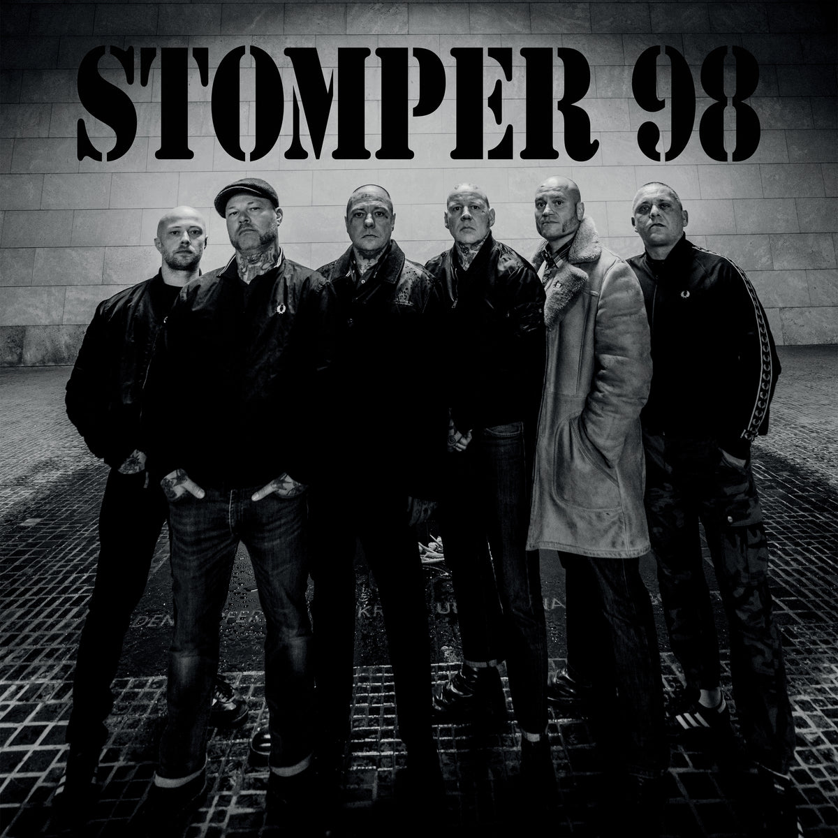 Six men with serious expressions and shaved heads stand together in a black and white photo. “Stomper 98” appears in bold above them, showcasing the brand’s iconic German Oi! punk rock roots against a stone wall backdrop.