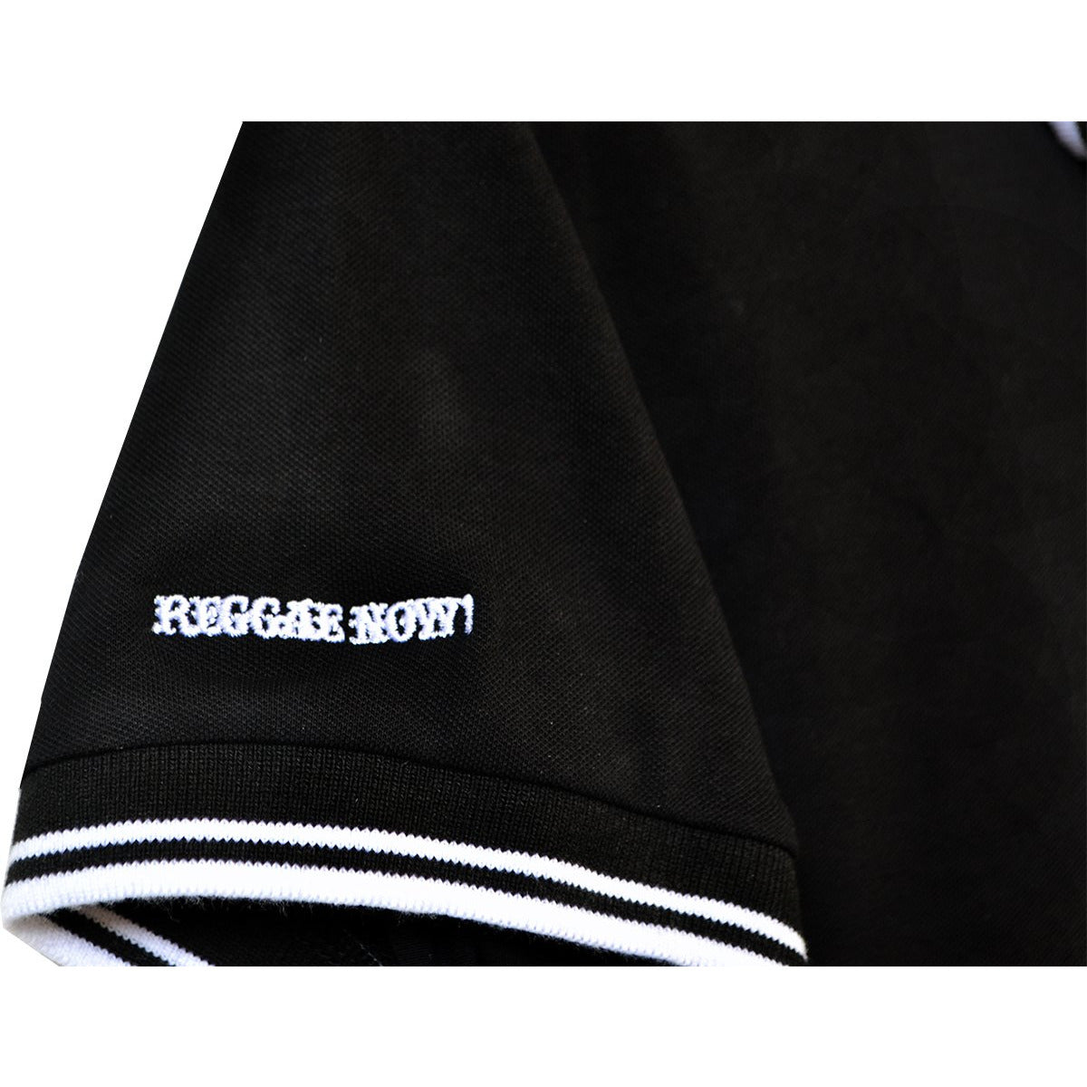 Close-up of a black The Aggrolites polo with white and black striped sleeve trim. REGGAE NOW! is embroidered in white near the hem—ideal for fans of classic reggae style and The Aggrolites.