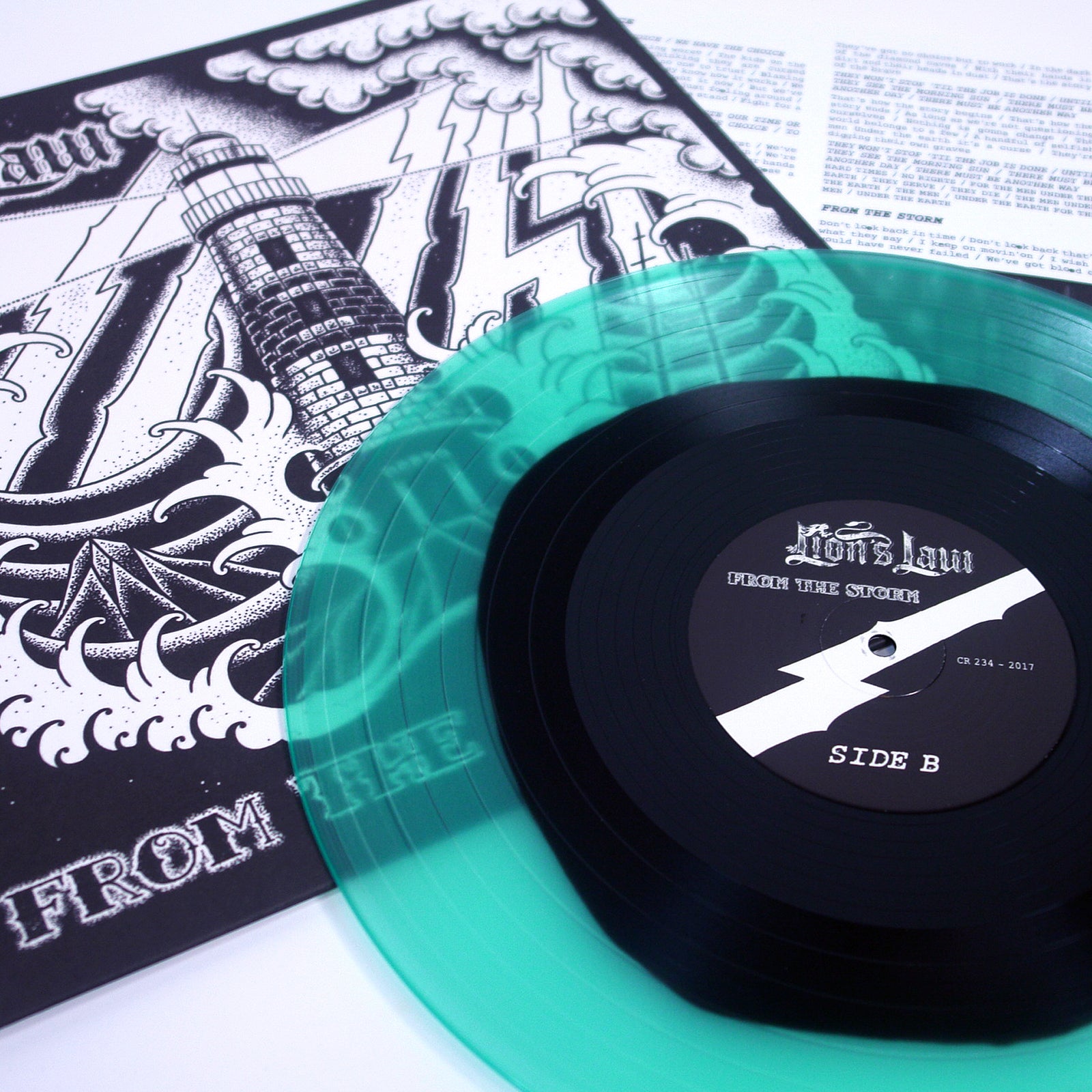 A translucent green and black vinyl record titled From the Storm by Lions Law is partially out of its illustrated cover with a lighthouse and waves, with a lyrics or info sheet from Lions Law visible in the background.