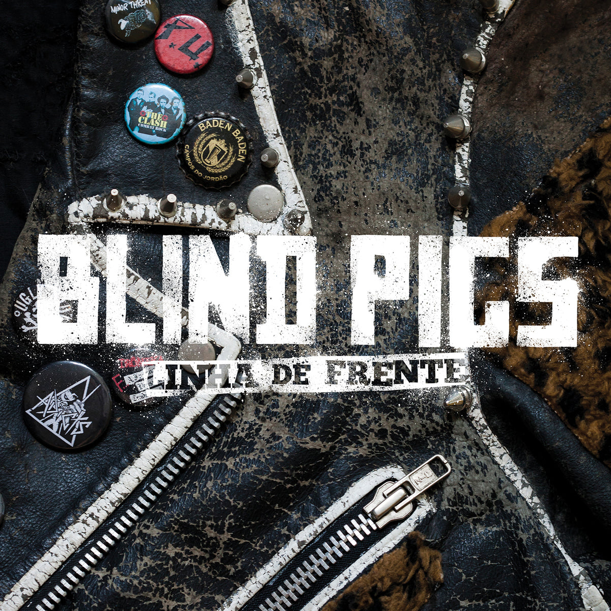 A close-up of a worn leather jacket with punk band pins and a white zipper. Bold white text reads Blind Pigs, iconic Brazilian streetpunk, with a smaller banner below saying Linha de Frente. Brand: Blind Pigs.
